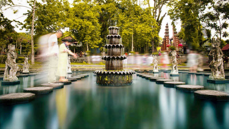 Visitors walking on stepping stones at Tirta Gangga Water Palace, Bali, with a tiered fountain and statues.