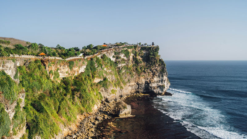 Scenic view of the Uluwatu cliff with lush greenery overlooking the Indian Ocean, a route leading to the sacred Uluwatu Temple in Bali.