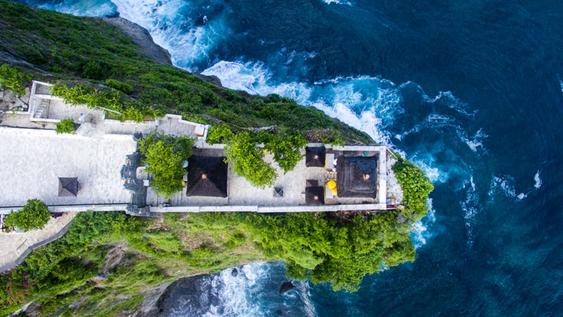 Arial view of the serene Uluwatu Temple perched on a lush cliff above frothy waves.