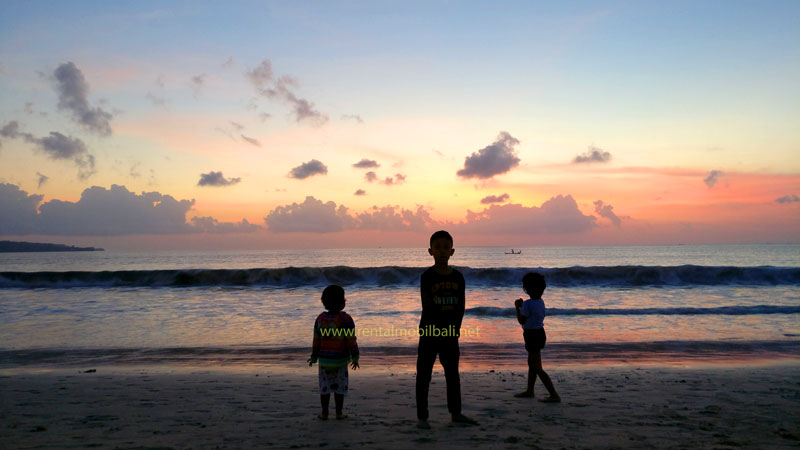 Three children joyfully playing on Jimbaran white sand beach during a vibrant sunset in Bali.