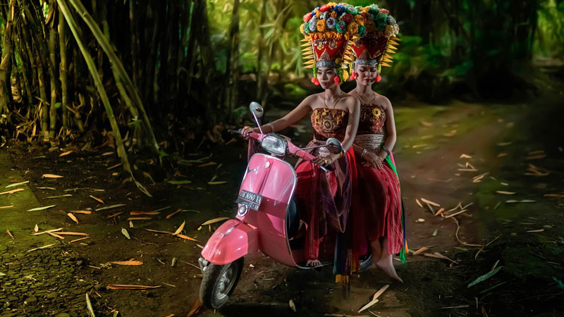 Two Balinese women in traditional attire posing with a colorful offering on a scooter.