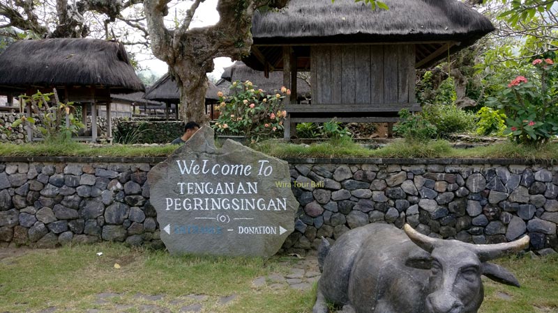 Entrance sign to Tenganan Pegringsingan Village in Bali with traditional Balinese huts and a stone bull sculpture.