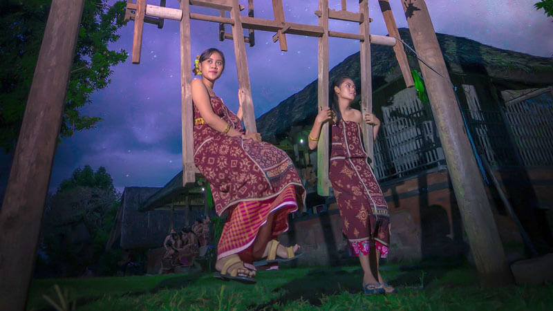 Young women in traditional Gringsing attire on swings in Tenganan Village at dusk.