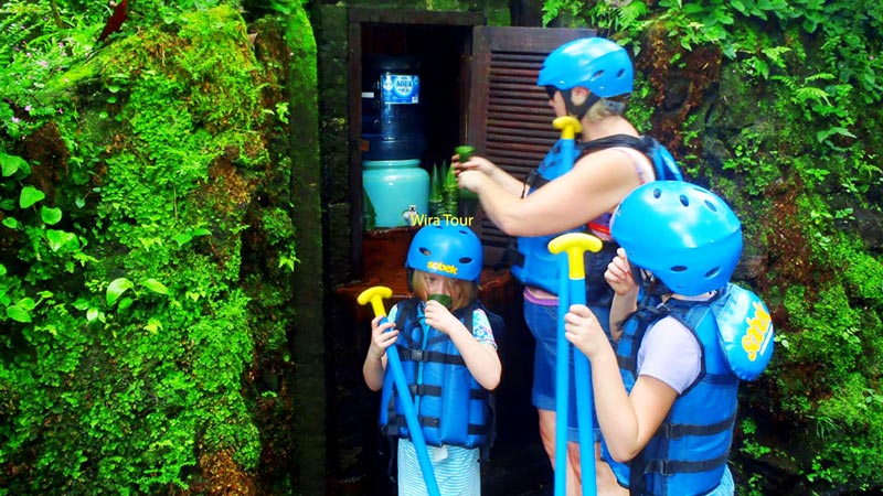 Family with two children geared up with safety helmets and life jackets for a river rafting adventure in Bali, showcasing Wira Tour branding.