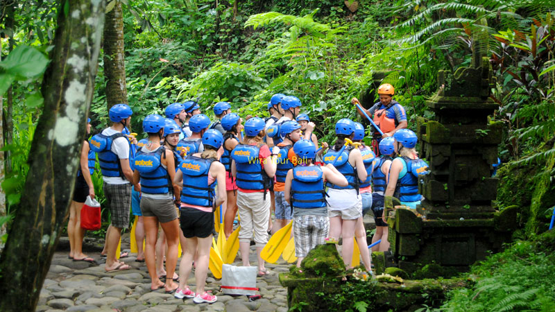 A group of rafters in blue and yellow gear attentively listening to a guide's safety briefing amidst Bali’s lush greenery.