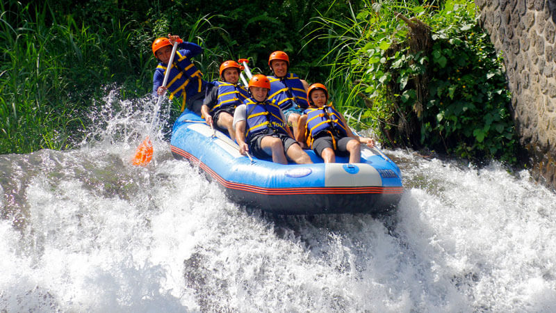 Group of adventurers-branded life jackets and helmets, navigating through a rapid in a raft in Bali's lush river surroundings.