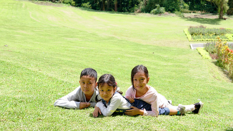 Three Balinese children smiling and laying on a vibrant green lawn in Bedugul.