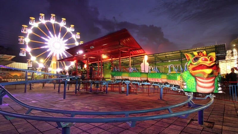 Brightly lit Ferris wheel at Krisna Funtastic Land in Bali during dusk with roller coaster track in the foreground