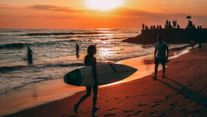 Surfer girl with a board at Bali sunset, with onlookers by a temple and others enjoying the beach waves