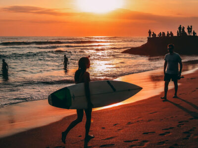 Surfer girl with a board at Bali sunset, with onlookers by a temple and others enjoying the beach waves