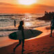 Surfer girl with a board at Bali sunset, with onlookers by a temple and others enjoying the beach waves