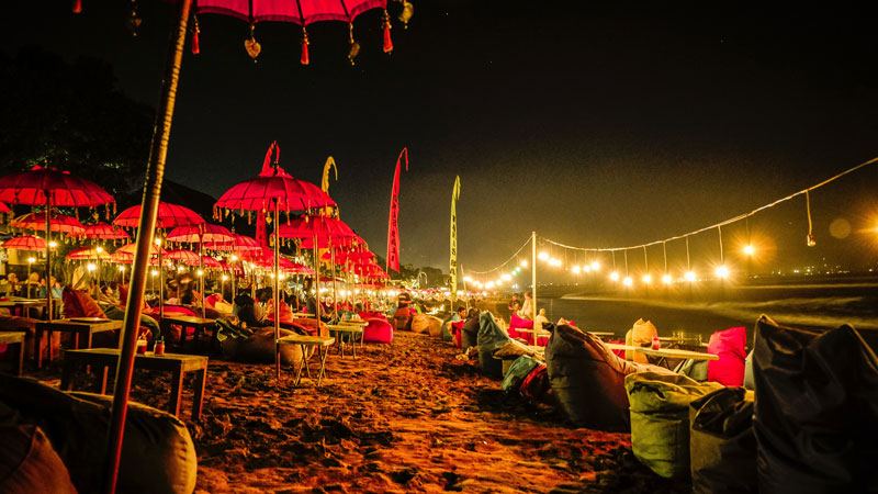 Nighttime beach setting with vibrant red umbrellas and visitors enjoying the Bali nightlife