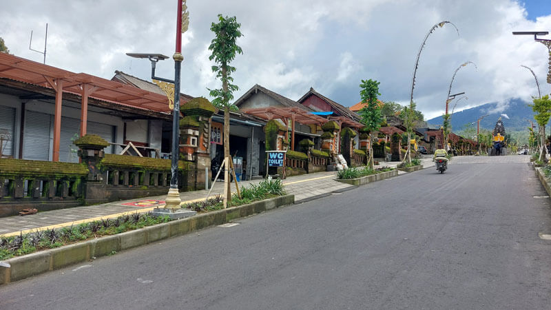 Approach road to holy site lined with traditional Balinese structures and lush greenery under a cloudy sky