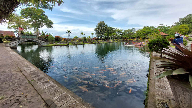 Visitors enjoying the serene atmosphere at Tirta Gangga Water Palace with a view of the ornate bridge over koi-filled ponds, set against lush Balinese gardens.