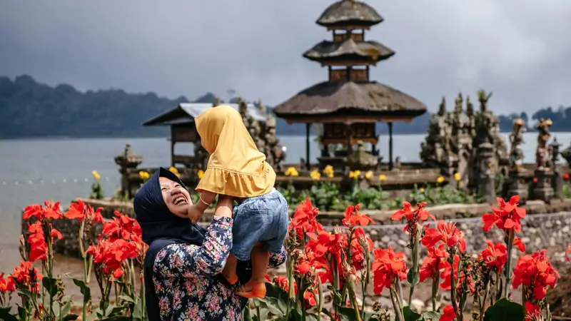 Smiling mother lifting child with Lake Beratan Temple in the background and red flowers in Bedugul Bali