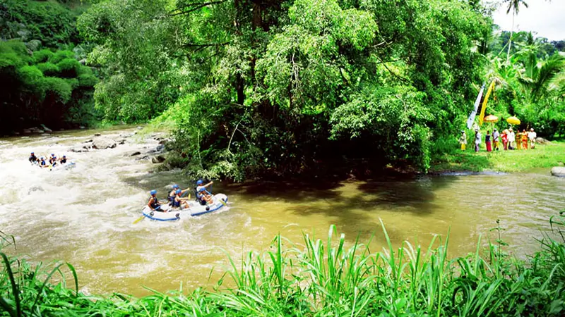 Rafts passing through jungle scenery with a Balinese ceremony visible on the riverbank.