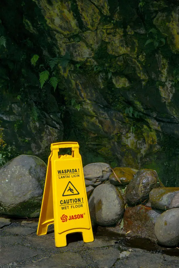 Yellow caution sign on a wet, mossy path after rain in Bali; slippery footing.