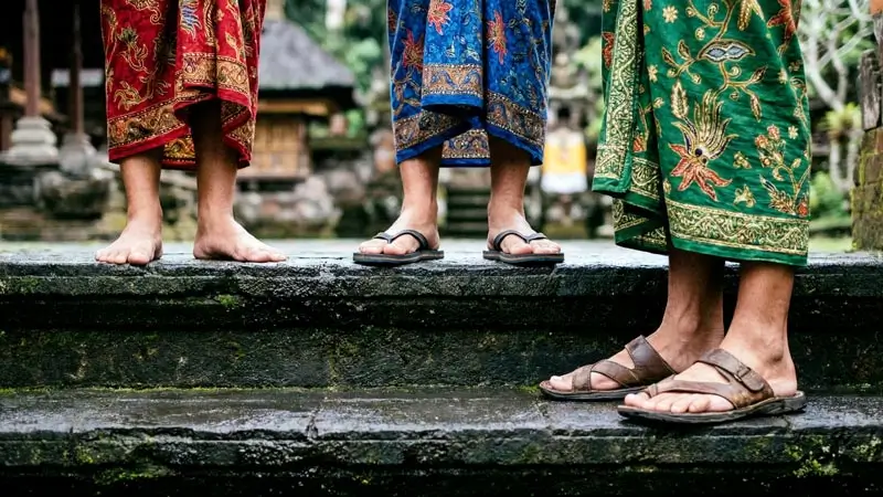 Feet in sandals and barefoot on wet Bali temple steps, wearing colourful sarongs for worship.