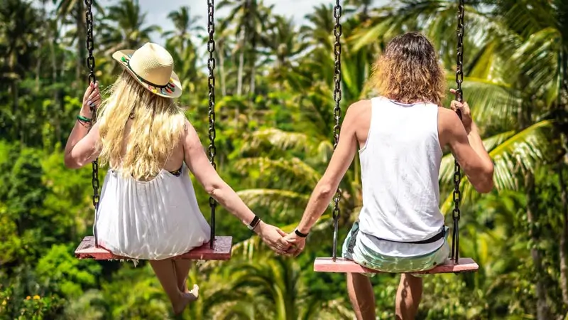 Couple holding hands on jungle swing in Ubud Bali with green palm trees behind
