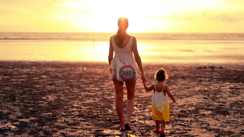 Mother and toddler walking on Bali beach at sunset.